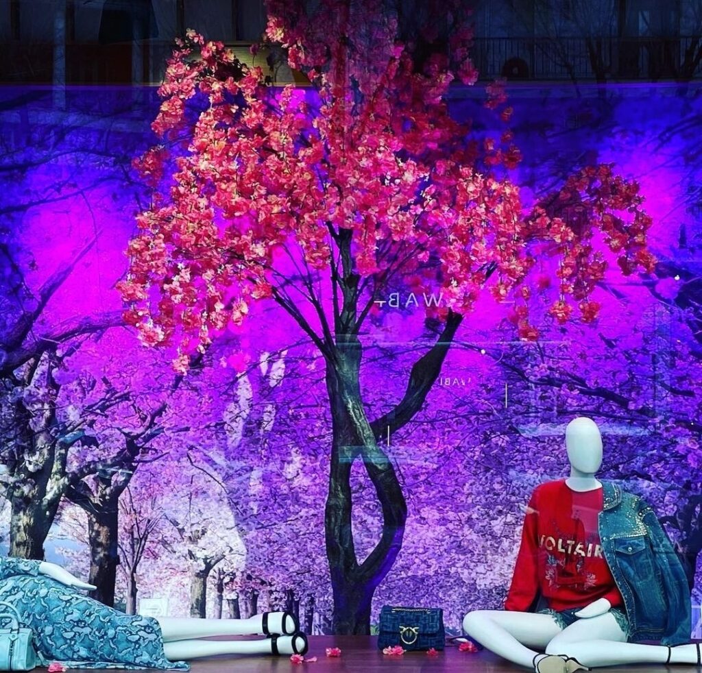 Storefront display featuring mannequins dressed in elegant clothing with a vibrant artificial tree and lush greenery, designed by Fairy Tale Green Walls in Limassol, Cyprus.